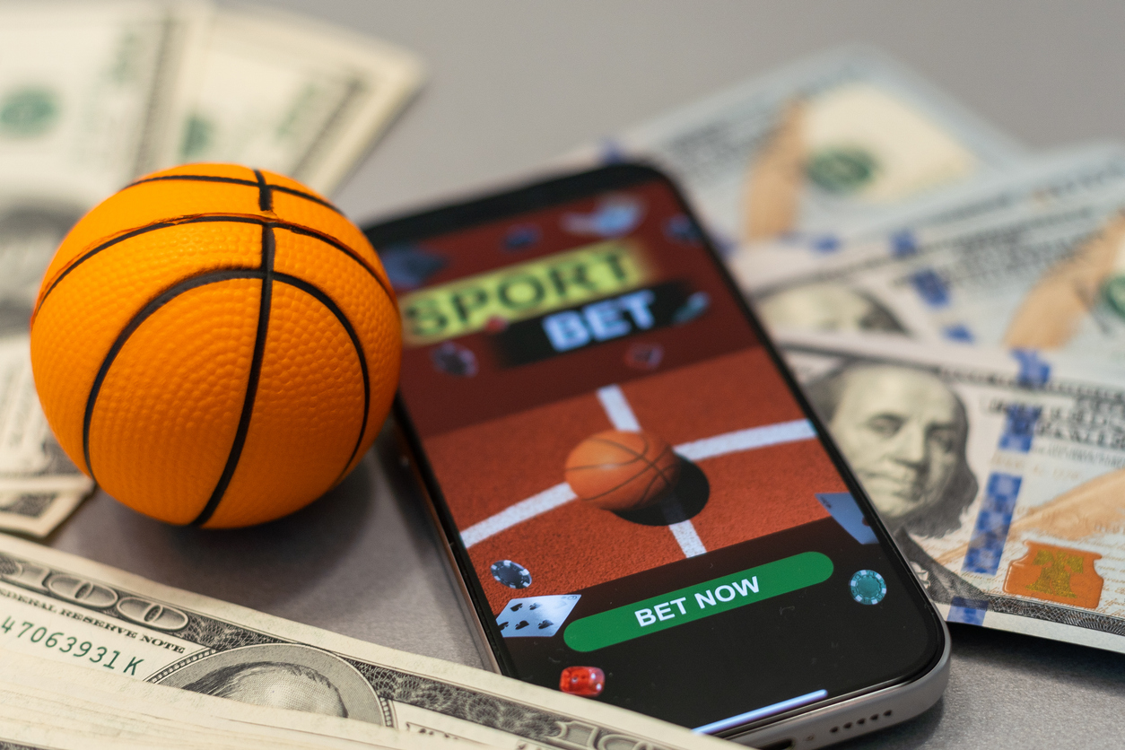 Sports betting app open on a phone beside cash and a basketball
